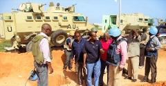 A group of people stand near an armored vehicle in a dusty area, some wearing helmets, likely engaged in peacekeeping or security operations.