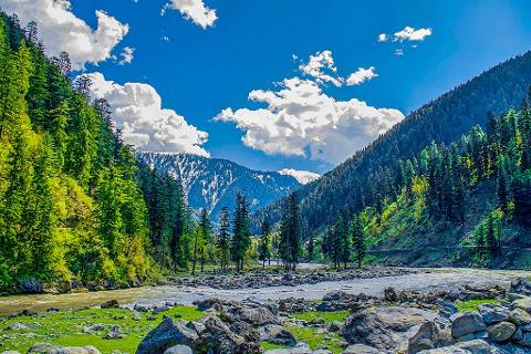 A scenic view of a river flowing through a valley with lush green mountains and snow-capped peaks under a blue sky with fluffy clouds.