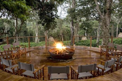 This image shows a fire pit on a wooden platform surrounded by chairs, set in a lush forest in the Odzala-Kokoua National Park.
