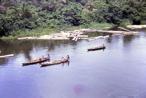 A river with rafts and three narrow boats with people carrying goods on the water, surrounded by lush green vegetation.