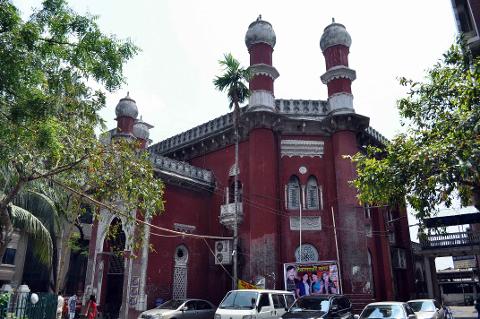The image shows the red brick building of the Dhaka High Court in Bangladesh, with parked cars in front.