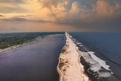 An aerial view shows a narrow sandy beach separating a dark river from the vast blue ocean under a dramatic, cloudy sky.
