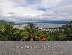 A scenic overlook with palm trees frames a view of the Rabaul Caldera, a town nestled along the bay, with ships in the distance and a cloudy sky above.