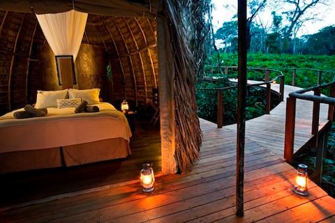 Here is the description of the image:A serene view of a luxurious jungle accommodation, featuring a cozy bed inside a thatched hut that opens onto a wooden boardwalk illuminated by lanterns amidst lush greenery.