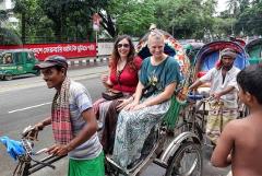 Two women are riding in a rickshaw being pulled by a man in a cap, while another man walks beside another rickshaw.