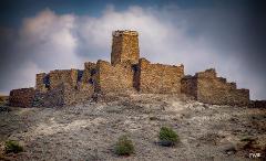 A stone fortress sits atop a barren hill under a cloudy sky, showcasing its weathered walls and central tower.