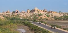 A road cuts through a landscape with a historic, dome-topped structure on a hill, featuring vehicles and a blend of arid terrain and green vegetation.