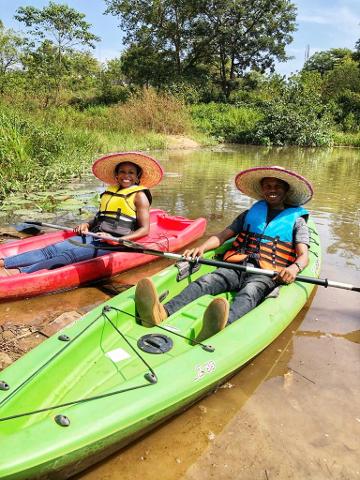 African Arts, Kayaking, and a Brewery Tour of Abuja Two people wearing life vests and straw hats are sitting in kayaks on a lake surrounded by lush greenery.