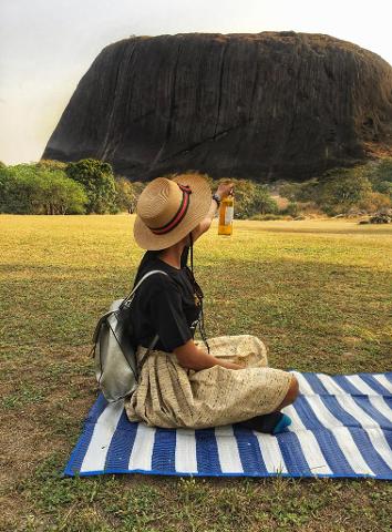 A woman wearing a hat and backpack sits on a striped blanket, holding up a bottle towards a large rock formation in the background.