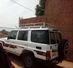 A white Land Cruiser with a roof rack and spare tire mounted on the back is parked next to a brown building and a metal fence.