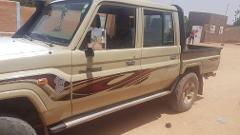 A tan Toyota Land Cruiser pickup truck with a snorkel and custom graphics is parked on a sandy surface in a rural area.