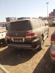 A dusty Lexus LX 570 is parked on a dry, grassy lot, possibly in a holding area or near a port, given the presence of shipping containers in the background.
