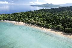 Aerial view of a tropical island featuring a lush green forest, a sandy beach with a wooden pier, and clear turquoise water with visible underwater patterns.