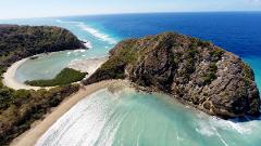 This aerial shot captures a stunning coastal landscape with turquoise waters, sandy beaches, and lush green hills meeting the horizon under a clear blue sky.