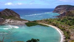 A scenic view of a tropical bay with turquoise water, white sand beach, and lush green hills under a bright blue sky.