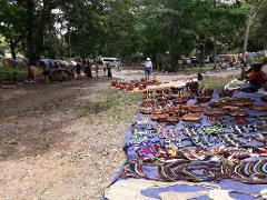 A bustling outdoor market in Port Moresby with colorful textiles and handcrafted baskets displayed on a large blue tarp, surrounded by people browsing and vendors.