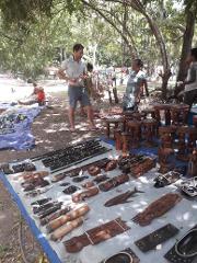 A tourist haggles with a local vendor displaying wooden crafts, including flutes and animal carvings, at an outdoor market in Port Moresby.
