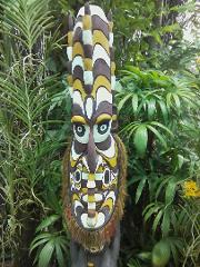 A tall, decorative mask stands among lush tropical foliage, featuring intricate patterns and a fringe of natural fibers.
