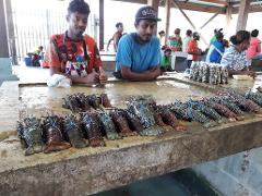 Two men are behind a table full of lobsters, likely at a market in Port Moresby. Other people are in the background.