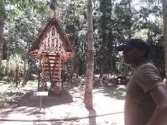 A man in a cap stands near a traditional structure with a straw roof and colorful designs in a park setting.