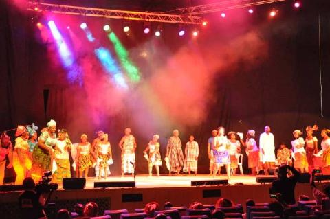 A stage performance featuring a group of people in colorful, traditional attire under bright stage lights and smoke, being watched by an audience.
