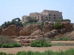 An ancient building atop a hill overlooks the crumbling ruins in the foreground, with lush greenery and a clear blue sky completing the scene.