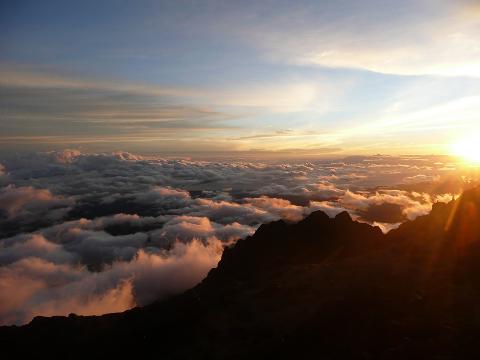 Sunrise over a sea of clouds, seen from a high mountain peak, casting a warm, golden light across the sky and mountain range.