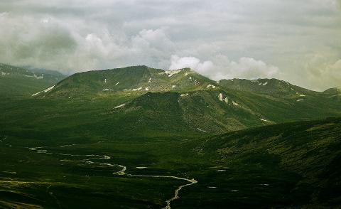 A winding river flows through a lush, green valley surrounded by rolling mountains under a cloudy sky.