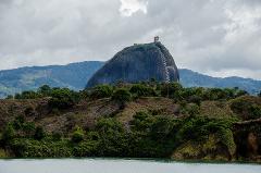 A large rock formation with a staircase and building at the top is surrounded by trees and water under a cloudy sky.