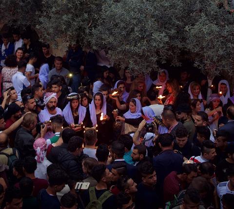 A crowd of people, many holding candles, gather under a tree, illuminated by the flickering flames and photographers capturing the scene.