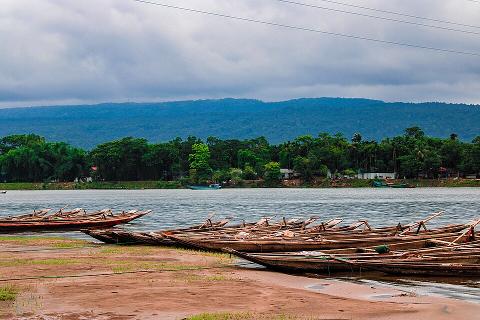 A serene view of a lake with several wooden boats lined up on the shore against a backdrop of lush green trees and distant mountains under a cloudy sky.