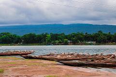 A serene view of a lake with several wooden boats lined up on the shore against a backdrop of lush green trees and distant mountains under a cloudy sky.