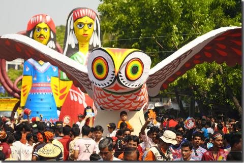 A crowd of people gathers around large, colorful, cartoonish figures, including an owl with outstretched wings and two human-like figures, likely as part of a cultural event or festival.
