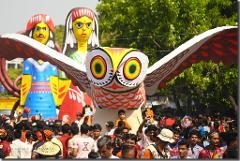 A crowd of people gathers around large, colorful, cartoonish figures, including an owl with outstretched wings and two human-like figures, likely as part of a cultural event or festival.