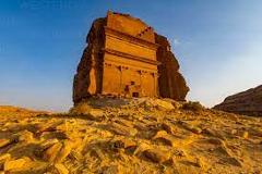 A monumental rock-cut tomb, likely from the Nabataean civilization, stands prominently against a blue sky in a desert landscape.