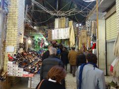 A bustling market scene with people walking through a narrow aisle lined with shoes, fabrics, and other goods hanging from the overhead structure.