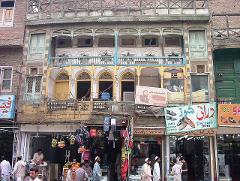An old, multi-story building with ornate balconies and decayed facade stands above street-level shops and vendors in a busy marketplace.
