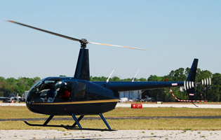 A dark blue Robinson R44 helicopter is sitting on a tarmac with its blades blurred from spinning.