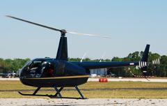 A dark blue Robinson R44 helicopter is sitting on a tarmac with its blades blurred from spinning.