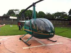 A green and brown Robinson R44 helicopter sits on a helipad, with people visible in the background on a grassy area.
