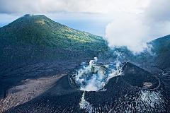 Aerial view of the Tavurvur volcano's caldera, steaming in the foreground, adjacent to a lush green mountain under a partly cloudy sky.