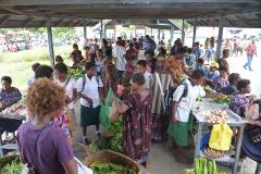 A busy market scene unfolds under a covered structure, with people browsing and purchasing vegetables, fruits, and other goods.