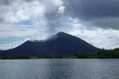 A dark volcano rises from the sea under a cloudy sky, with a line of greenery at its base.