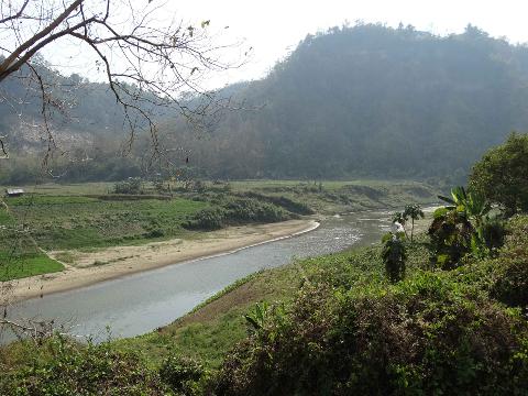 A scenic view of a river winding through a valley, flanked by lush greenery and a backdrop of hills under a hazy sky.