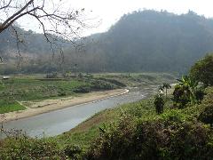 A scenic view of a river winding through a valley, flanked by lush greenery and a backdrop of hills under a hazy sky.