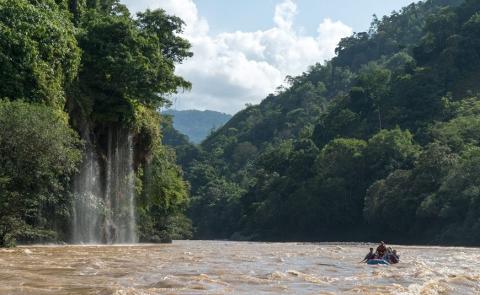 A group of people in a raft are navigating a river with muddy water near a waterfall, surrounded by lush green vegetation and mountains in the background.