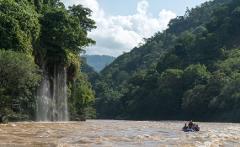 A group of people in a raft are navigating a river with muddy water near a waterfall, surrounded by lush green vegetation and mountains in the background.