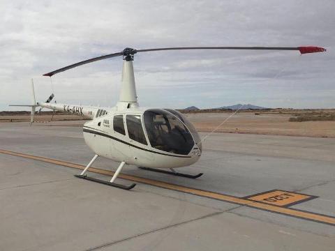 A white Robinson R66 Turbine helicopter sits on a tarmac with mountains visible in the background under a cloudy sky.