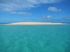 A sandbar is surrounded by clear turquoise water under a blue sky with scattered clouds.