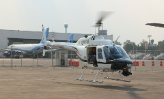 A white helicopter with a man inside hovers on a tarmac, with an airplane, air traffic control tower, and stairs in the background, presumably related to a tour of the Teotihuacan Pyramids.